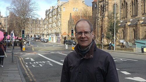 Neil Ross standing in front of Holy Corner, a junction in Morningside, with churches and tenements in the background. The sky is blue.