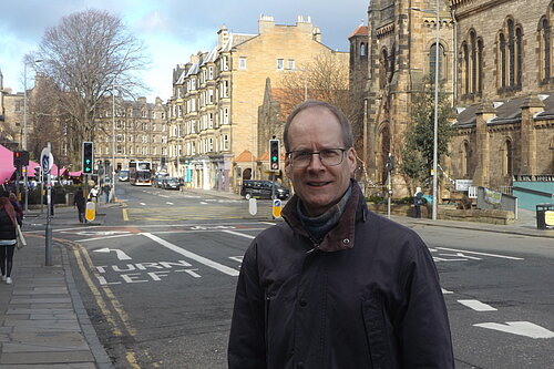 Neil Ross standing at Holy Corner, Morningside. A junction, churches, tenements and a blue sky are in the background.