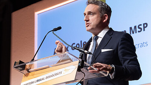 Alex Cole-Hamilton speaking at a podium with the words "Scottish Liberal Democrats" on it